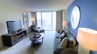 This well-appointed single-floor residence offers a refined living experience defined by thoughtful design and an abundance of natural light. The living area serves as the heart of the space, anchored by expansive floor-to-ceiling windows that frame sweeping views of the surrounding cityscape and greenery beyond. A sliding glass door opens to the outdoors, further connecting the interior to its elevated surroundings.

The room is elegantly furnished with a plush grey sofa adorned with decorative pillows, a matching chaise lounge, and a sleek oval coffee table — creating a comfortable and cohesive seating arrangement. A bold blue accent wall adds a striking pop of color and visual depth, complemented by a large circular mirror that enhances the sense of openness. Warm ambient lighting from stylish table lamps contributes to an inviting atmosphere during evening hours.

Soft wall-to-wall carpeting runs throughout the living space, adding warmth underfoot, while neutral tones on the walls and ceiling maintain a clean, modern aesthetic. A media console along one wall provides functional storage and display space. Overall, the space strikes a balance between contemporary style and comfortable livability, making it an ideal retreat with a stunning elevated urban outlook.