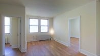 This single-floor space offers a clean, understated aesthetic defined by light hardwood flooring and soft, neutral-toned walls that create a bright and welcoming atmosphere. The main living area is anchored by two side-by-side windows that allow generous natural light to fill the room, illuminating the warm tones of the wood floor throughout. A classic white radiator sits beneath the windows, indicating a traditional heating system consistent with established building construction. The white baseboards and door trim provide a crisp contrast against the pale walls, adding a sense of polish to the otherwise minimalist interior. An open doorway on the right side of the room leads seamlessly into an adjoining space, suggesting a functional, connected floor plan. The absence of interior clutter emphasizes the roominess of the layout and the versatility of the space for various furniture arrangements. Overall, the space conveys a simple, practical character with a neutral palette that serves as an adaptable canvas for personalization.