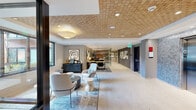 Located at 6312 9th Ave NE in Seattle's vibrant Ravenna neighborhood, this thoughtfully designed lobby space offers a refined and welcoming environment that sets the tone for the building it serves. The single-floor layout extends in a long, open configuration, maximizing flow and accessibility throughout.

A defining architectural feature is the geometric, woven-pattern wood ceiling, which adds warmth and visual texture throughout the space. Recessed lighting complements the natural tones of the ceiling, ensuring consistent, well-distributed illumination. The flooring consists of polished concrete-style material, lending a clean, contemporary feel underfoot.

Upon entry through sleek, black-framed glass doors, visitors are greeted by a built-in upholstered bench with channel-tufted leather detailing, positioned beneath a striking large-scale botanical wall sculpture in vibrant blues and greens—serving as a central artistic focal point. Adjacent to the entry, a comfortable seating area features modern lounge chairs, layered area rugs, and accent tables, creating an inviting space for informal gathering.

Further into the space, a workstation area provides functional desk seating, while a stone-clad elevator bank with stainless steel doors anchors the far end. Textured stone wall panels introduce additional material contrast. A curated gallery wall with framed artwork adds visual interest along the corridor, contributing to an overall atmosphere that is polished, contemporary, and refined.