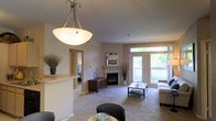 This single-floor residence is designed with an open, flowing layout that connects the dining and living areas seamlessly. The living room serves as the centerpiece of the space, anchored by a classic corner fireplace with a light-toned surround and a decorative mirror mounted above. A dark wood media console sits adjacent to the fireplace, providing functional storage while complementing the warm, neutral color palette of the walls and plush carpeted flooring throughout.

Large windows and a glass-paneled exterior door flood the space with natural light and offer views of a greenery-lined outdoor balcony, creating a bright and inviting atmosphere. A comfortable seating arrangement surrounds a dark wood coffee table, ideal for both relaxation and entertaining.

The dining area is defined by an elegant pendant light fixture with a frosted bowl shade, positioned near a raised breakfast bar that acts as a subtle divider between the kitchen and living spaces. Decorative touches such as potted plants and sculptural accents add warmth and personality to the interior. A glimpse into an adjacent room suggests additional private living quarters beyond the main living area.