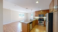 This single-floor residence offers a thoughtfully designed open-concept layout that connects the kitchen and living areas seamlessly. The kitchen is a functional centerpiece, featuring warm honey-toned cabinetry, granite countertops, a large center island with a built-in sink and dishwasher, and a full suite of stainless steel appliances including a gas range, over-the-range microwave, and refrigerator. Pendant lighting above the island adds a refined touch, while recessed lighting throughout ensures consistent illumination. The open living area is generously proportioned, bathed in natural light from multiple windows that offer views of surrounding greenery. Light hardwood-style flooring extends throughout the main living spaces, contributing to a bright and cohesive aesthetic. A hallway connects the living spaces to the private areas of the residence, where carpeted bedrooms provide a comfortable and quiet retreat. Each bedroom features neutral-toned walls and windows with blinds. The bathroom is equipped with a soaking tub and shower combination featuring built-in shelving, tile surround, and tile flooring, all finished in crisp white tones. Clean lines, neutral color palettes, and consistent finishes throughout give the space a modern, move-in-ready character.