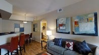 This single-story space offers a thoughtfully designed open floor plan that seamlessly connects the living and dining areas. The living room serves as the central hub, featuring warm hardwood-style flooring that runs throughout, neutral-toned walls, and ample ceiling height that contributes to an airy feel. A comfortable gray sofa anchored by a sleek glass coffee table takes center stage, complemented by vibrant abstract artwork that adds pops of color and personality to the walls.

The dining area sits adjacent to the living space, furnished with a compact table and red dining chairs beneath a contemporary pendant light, creating a defined yet open zone for meals. An arched doorway leads to a bathroom, which is warmly lit and provides a welcoming transition between spaces. The bathroom is visible from the living area and appears bright and functional.

Natural and artificial lighting work together throughout the space, with ceiling vents and recessed fixtures ensuring comfort. The cohesive color palette of warm neutrals, accented by bold blues and reds in furnishings and artwork, gives the space a modern yet inviting ambiance suitable for everyday living.