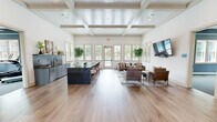 Located on Denali Drive in Mountain Park, Georgia, this thoughtfully designed single-floor community space blends classic architectural detail with contemporary style. The interior is defined by striking coffered and beadboard ceilings that add considerable visual height and elegance throughout the main gathering area. Wide-plank hardwood flooring runs continuously across the space, lending warmth and cohesion to every zone.

The main hall opens into a generously proportioned lounge area featuring multiple seating arrangements with leather and upholstered chairs, area rugs, and a wall-mounted flat-screen TV — ideal for both casual gatherings and organized events. An abundance of natural light pours in through a wide bank of double-hung windows and French doors along the far wall, offering views of the surrounding greenery.

A sleek wet bar and prep area with blue-gray cabinetry, subway tile backsplash, and a dark granite-topped island with bar seating anchors one side of the room, making it well-suited for entertaining. The entrance area features a modern globe chandelier, blue-gray board-and-batten accent walls, a water station, and an adjacent glass-walled conference or meeting room. A secondary lounge nook with a textured accent wall and decorative artwork provides a more intimate retreat within the space.