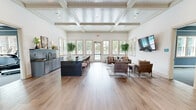 Located on Denali Drive in Mountain Park, Georgia, this thoughtfully designed single-floor community space blends classic architectural detail with contemporary style. The interior is defined by striking coffered and beadboard ceilings that add considerable visual height and elegance throughout the main gathering area. Wide-plank hardwood flooring runs continuously across the space, lending warmth and cohesion to every zone.

The main hall opens into a generously proportioned lounge area featuring multiple seating arrangements with leather and upholstered chairs, area rugs, and a wall-mounted flat-screen TV — ideal for both casual gatherings and organized events. An abundance of natural light pours in through a wide bank of double-hung windows and French doors along the far wall, offering views of the surrounding greenery.

A sleek wet bar and prep area with blue-gray cabinetry, subway tile backsplash, and a dark granite-topped island with bar seating anchors one side of the room, making it well-suited for entertaining. The entrance area features a modern globe chandelier, blue-gray board-and-batten accent walls, a water station, and an adjacent glass-walled conference or meeting room. A secondary lounge nook with a textured accent wall and decorative artwork provides a more intimate retreat within the space.