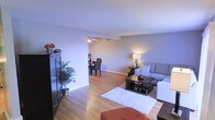 This 2-floor residence offers a thoughtfully designed layout that balances comfort and functionality. The main living area is an open-concept space where the living room flows naturally into the dining area, creating a cohesive and welcoming environment. Warm hardwood flooring extends throughout, complementing the soft, neutral wall tones that lend the space a calm and airy ambiance.

The living room is generously sized, offering ample room for seating arrangements centered around a dark wood coffee table. A plush area rug anchors the space, while a large sectional sofa provides comfortable seating. A dark wood media cabinet stands along one wall, adding both storage and visual contrast to the light-toned surroundings.

Adjacent to the living room, the dining area is defined by its ceiling fan overhead and charming floral centerpiece, accommodating a dark wood dining set suitable for everyday meals and entertaining. Warm lamp lighting ties the two areas together, contributing to the inviting atmosphere throughout.

The open sightlines between rooms enhance the sense of spaciousness, while clean white baseboards and smooth ceilings give the interior a polished, well-maintained appearance. With two floors, this home offers additional living space beyond what is immediately visible, providing versatility for a variety of lifestyle needs.