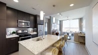 This single-floor space showcases a sleek, open-concept layout that seamlessly connects the kitchen and living areas. The kitchen is a standout feature, anchored by a generous island with a light-veined stone countertop that doubles as a breakfast bar with stylish gold wire seating. Dark espresso cabinetry contrasts elegantly with the light countertops, while stainless steel appliances — including a gas range, refrigerator, and over-the-range microwave — add a polished, professional feel. Cylindrical pendant lights suspended above the island provide warm, focused illumination.

The space transitions naturally into the living area, which benefits from floor-to-ceiling windows that flood the interior with natural light and offer open views beyond. Wood-toned flooring runs throughout, reinforcing the warm, contemporary aesthetic. A ceiling fan in the living area adds a functional element while maintaining the modern design language. Track lighting and recessed fixtures ensure the entire space is well-lit at any time of day. The overall ambiance is clean, sophisticated, and thoughtfully designed for both everyday living and entertaining.