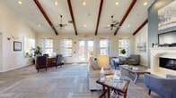 Located at 513 Dodson Rd in Rogers, Arkansas, this single-story community clubhouse offers a well-appointed gathering space designed for both comfort and functionality. The centerpiece of the space is a generously sized lounge area distinguished by soaring vaulted ceilings adorned with rich, dark-stained exposed wood beams and dual ceiling fans. Large windows dressed with patterned curtains flood the room with abundant natural light, while glass French doors provide direct access to an outdoor area. Comfortable seating arrangements, including sofas and accent chairs, are thoughtfully arranged throughout the lounge, which also features a white-mantled fireplace accented by a bold slate-blue accent wall and a wall-mounted television — creating a warm, inviting focal point. The kitchen area is fully equipped with white cabinetry, granite countertops, stainless steel appliances including a French door refrigerator and microwave, and a rustic wood-paneled island with bar seating for casual dining. Adjacent to the kitchen, a dedicated workspace with built-in desk and computer provides a convenient area for residents. Neutral carpet tile flooring and recessed lighting throughout the space contribute to a clean, cohesive aesthetic that is both practical and welcoming.