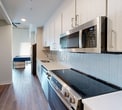 This efficiently designed 280 square foot studio unit is laid out across a single floor, making smart use of every square foot. The open-concept living and sleeping area features warm hardwood-style flooring, high ceilings, and a wall-mounted TV niche, creating a modern and functional living space. A ceiling fan provides additional comfort overhead. The galley-style kitchen is well-appointed with stainless steel appliances including a refrigerator, electric range, microwave, and dishwasher, complemented by white cabinetry with sleek hardware, a light quartz countertop, an undermount sink, and a subtle tile backsplash. Adjacent to the kitchen, a dedicated built-in shelving and storage unit offers practical organizational space. The entryway features a bold navy front door and high ceilings that enhance the sense of openness. The bathroom includes a glass-door shower with tiled surround, a toilet, and consistent hardwood-style flooring that ties the space together. Throughout the unit, a neutral white and warm wood color palette contributes to a clean, contemporary aesthetic well-suited for efficient urban living.