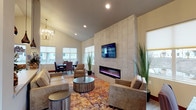 Located at 574 West Mogul Loop in Hayden, Idaho, this single-story community clubhouse serves as the welcome center for the Villas at Hayden. The space is thoughtfully designed to combine comfort and functionality in a modern, inviting atmosphere.

Upon entry, a bright foyer greets visitors with soaring vaulted ceilings, light wood-tone flooring, and a pair of upholstered accent chairs positioned beside the branded Villas at Hayden logo window. Pendant lighting and a statement chandelier add warm, layered illumination throughout.

The main lounge area is the centerpiece of the clubhouse, anchored by a dramatic floor-to-ceiling light-tiled accent wall that houses a wide linear electric fireplace and a wall-mounted flat-screen television. Comfortable neutral-toned sofas and armchairs are arranged around a rustic round coffee table, all set atop a vibrant, multi-colored area rug that adds energy and warmth to the space. Ample natural light streams in through multiple large windows fitted with roller shades.

Adjacent to the lounge, a casual dining area features a solid wood table with upholstered chairs, ideal for resident gatherings. A reception counter with pendant lighting connects to an administrative corridor leading further into the building. Together, these spaces create a welcoming, well-appointed community hub.