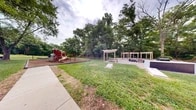 Located at 8818 Blairwood Road in Nottingham, Maryland, this outdoor community recreation space offers a well-rounded array of amenities designed for residents of all ages. The grounds are anchored by two distinct playground areas, each set on rubberized mulch surfaces for safety. One features a multi-level structure with climbing walls, spiral and straight slides, and interactive elements, while a second, smaller unit provides additional play options for younger children. A wooden pergola structure spans a paved patio area, offering shaded seating with benches arranged beneath its open-beam canopy — an ideal spot for gatherings and outdoor leisure. A cornhole board nearby adds to the recreational appeal. Expansive, well-maintained green lawns provide generous open space for recreation and relaxation. Concrete walkways connect the various areas, lined with decorative wooden archway structures that add architectural character to the grounds. Mature trees frame the perimeter, creating a natural, park-like atmosphere, while a split-rail fence along one boundary borders a lush woodland backdrop. The neighboring brick residential buildings provide a cohesive community context, making this a vibrant and functional outdoor hub for the Nottingham community.