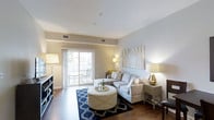 This single-floor residence offers a thoughtfully designed living space that balances modern aesthetics with everyday comfort. The living room serves as the heart of the home, featuring warm hardwood flooring, soft neutral-toned walls, and a clean, open layout that creates an inviting atmosphere. A large sliding glass door floods the room with natural light and provides seamless access to a private outdoor patio, where outdoor seating can be glimpsed beyond the sheer curtains. The living area is well-proportioned, accommodating a plush sectional sofa, a tufted round ottoman centered on a bold navy geometric area rug, and coordinating accent furniture. Warm lamp lighting complements the natural light, adding to the welcoming ambiance throughout the day and evening. Wall art featuring a nature-inspired print adds a subtle artistic touch to the space. Built-in shelving along one wall provides both storage and display opportunities, adding functional character to the room. The overall design presents a harmonious blend of contemporary furnishings and neutral finishes, creating a space that feels both polished and livable.