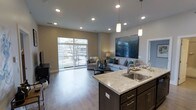 This single-floor residence showcases a thoughtfully designed open-concept layout that seamlessly integrates the kitchen and living areas into one cohesive, flowing space. The kitchen features a substantial island with light stone countertops, dark espresso cabinetry with brushed metal hardware, and a stainless steel sink. Pendant lighting above the island adds a stylish focal point, complementing the recessed lighting that illuminates the entire space.

The living area benefits from an abundance of natural light streaming through large floor-to-ceiling windows, which also provide direct access to a private balcony overlooking outdoor greenery. The neutral, warm-toned walls and hardwood-style plank flooring create a clean, modern aesthetic throughout. A large statement artwork in deep blues and teals anchors the living area, adding a vibrant contrast to the otherwise neutral palette.

The open layout promotes a sense of spaciousness, making the area feel expansive and airy. The overall ambiance is contemporary and refined, with a well-balanced combination of functional design elements and modern finishes that create an inviting and comfortable living environment.