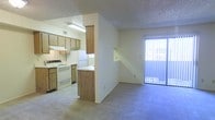 This single-floor residence offers a practical and comfortable layout designed to maximize functionality. The kitchen is well-appointed with ample wooden cabinetry providing generous storage, a full suite of white appliances including a range and refrigerator, and a built-in ventilation hood. A half-wall with a wood-panel breakfast bar visually separates the kitchen from the adjoining living space while maintaining an open, connected feel throughout.

The living area is finished with wall-to-wall carpeting in a neutral tone, creating a warm and comfortable atmosphere. A textured ceiling runs throughout the unit, adding subtle dimension to the interior. Natural light filters into the living space through a large sliding glass door dressed with vertical blinds, which opens to an outdoor area and provides a bright, airy ambiance.

The open passage between the kitchen and living room creates a seamless flow ideal for everyday living. The neutral color palette of cream and beige tones throughout the walls, flooring, and cabinetry lends the space a cohesive and understated aesthetic. A small niche in the wall near the living area offers an additional decorative or functional detail. Overall, the layout is efficient and well-organized, making it well-suited for comfortable day-to-day living.