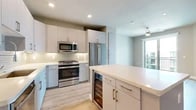 This thoughtfully designed one-bedroom, one-bathroom residence offers a contemporary living experience with a clean, modern aesthetic throughout. The kitchen is a standout feature, showcasing sleek flat-front cabinetry in soft gray and white tones, stainless steel appliances including a gas range, built-in microwave, French door refrigerator, and dishwasher. A subway tile backsplash adds a refined touch, while the central island with quartz countertops and an integrated wine cooler makes the space ideal for both cooking and entertaining. Light hardwood-style flooring flows through the kitchen and living areas, complementing the bright, neutral color palette. The bedroom is carpeted for comfort, finished with warm taupe walls, a ceiling fan, and a window with views of a private balcony, allowing natural light to fill the room. A walk-in closet adjoins the bedroom, offering built-in shelving and ample storage. The bathroom features a white quartz vanity with generous drawer storage, a large mirror, and a glass-enclosed walk-in shower with clean tile finishes. Recessed lighting throughout and a cohesive modern design make this a well-structured and functional living space.