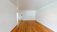 This single-floor, 1-bedroom, 1-bathroom unit offers a thoughtfully organized layout across five distinct spaces: a living room, bedroom, kitchen, bathroom, and hallway. The living room is a standout feature, offering generous proportions with warm-toned hardwood flooring throughout, crisp white walls, and a wall-mounted heating unit. Natural light filters in through a sliding glass door framed by curtains, opening to an outdoor view. The entryway leads directly into this open, airy living space, reinforcing a welcoming flow throughout the unit. The kitchen is bright and functional, equipped with a stainless steel gas range and white cabinetry. A window above the counter area looks out to an outdoor seating area, providing pleasant natural light. The bedroom is a comfortable size, featuring the same rich hardwood flooring, a ceiling fan, a natural light-filled window, and a mirrored sliding closet with built-in upper storage cabinetry. The bathroom is clean and modern, with a white vanity cabinet, an integrated sink with chrome fixtures, a bathtub, a wall-mounted mirror, and a window that provides natural ventilation. Gray wood-look flooring grounds the space. Overall, the unit presents a cohesive, light-filled aesthetic defined by its warm hardwood floors and clean white finishes throughout.