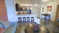 This single-story residence showcases an open-concept layout that integrates the kitchen, dining, and living areas into one cohesive and functional space. The kitchen is equipped with dark espresso cabinetry, a black refrigerator, and ample counter space, complemented by pendant lighting that hangs elegantly above the extended breakfast bar. The bar-height counter comfortably accommodates four cushioned swivel stools, making it an ideal spot for casual dining or entertaining. Track lighting and flush-mount ceiling fixtures provide consistent illumination throughout the space. Polished concrete floors extend across the entire living area, offering a sleek, low-maintenance surface that adds an industrial-modern character to the interior. A defined dining nook is visible toward the rear, furnished with a tall table and seating, while the living area anchors the foreground with a decorative round coffee table set atop a patterned area rug. A paneled door provides access to an adjoining room, and framed artwork on the walls adds a finishing touch to the well-appointed interior.