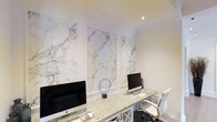 This sophisticated single-floor space is defined by its elegant, high-end design details and a cohesive white-and-stone aesthetic. The standout feature of the workspace is the accent wall, which showcases a series of large, framed marble panels with sweeping grey and gold veining set against crisp white walls — a bold yet refined design choice that immediately draws the eye.

The work surface runs generously along the wall, finished in a speckled granite that complements the marble panels above. The desk area is designed to accommodate multiple workstations, providing ample surface area for productivity. White cabinetry with classic cross-detail paneling lines the lower half of the workspace, adding a traditional architectural touch that balances the contemporary use of stone.

Recessed ceiling lighting provides focused illumination throughout, ensuring a well-lit and functional environment without disrupting the clean lines of the interior. A white ergonomic chair completes the workstation setup, reinforcing the consistent, monochromatic design palette.

Overall, the space communicates a sense of understated luxury — carefully curated with premium materials and a disciplined color scheme that is both visually impressive and highly functional.
