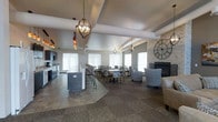This single-floor space is designed as a multifunctional communal gathering area, thoughtfully arranged to accommodate a variety of social settings. The open-concept layout seamlessly connects a kitchenette and bar area to a casual dining zone and a relaxed lounge corner, creating a cohesive and inviting environment throughout.

The ceiling features exposed beams and is adorned with a collection of stylish globe and cage-style pendant light fixtures, adding a warm, contemporary character to the space. The flooring transitions between stone tile and patterned carpet, visually defining the different functional zones while maintaining a unified aesthetic.

Along one side, a kitchenette with dark cabinetry and stainless steel appliances is paired with a raised bar counter and bar-height seating. The central dining area accommodates multiple tables and chairs, suitable for casual meals or group activities. Large windows dressed with white blinds allow natural light to fill the room.

On the far wall, a stone-surround fireplace anchors the lounge area, where comfortable upholstered chairs provide a cozy retreat. A large decorative clock mounted on the stone wall adds a distinctive focal point, completing the space's warm and welcoming ambiance.