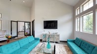 The Media Lounge is a thoughtfully designed communal entertainment space that combines bold style with comfortable functionality. The room immediately draws the eye upward with its dramatically high vaulted ceilings, which lend an open, airy quality to the space and amplify the sense of volume throughout. Crisp white walls serve as a clean, neutral backdrop that highlights the room's vivid design choices.

At the heart of the lounge is a large wall-mounted flat-screen television, positioned above a decorative mirrored console cabinet that adds a touch of elegance. Two striking teal tufted sofas face each other across a sleek glass-topped coffee table, anchored by a plush ivory shag area rug that adds warmth and texture underfoot. The bold turquoise seating provides a vibrant pop of color that energizes the space and creates a welcoming, social atmosphere.

A glimpse through the doorway reveals an adjacent area with additional amenities, suggesting the lounge is part of a broader community facility. The dark-framed doorway and dark baseboard trim provide sharp contrast against the light walls, reinforcing the modern aesthetic. Overall, the Media Lounge offers a polished, inviting environment well-suited for relaxation, socializing, and shared entertainment experiences.