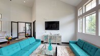 The Media Lounge is a thoughtfully designed communal entertainment space that combines bold style with comfortable functionality. The room immediately draws the eye upward with its dramatically high vaulted ceilings, which lend an open, airy quality to the space and amplify the sense of volume throughout. Crisp white walls serve as a clean, neutral backdrop that highlights the room's vivid design choices.

At the heart of the lounge is a large wall-mounted flat-screen television, positioned above a decorative mirrored console cabinet that adds a touch of elegance. Two striking teal tufted sofas face each other across a sleek glass-topped coffee table, anchored by a plush ivory shag area rug that adds warmth and texture underfoot. The bold turquoise seating provides a vibrant pop of color that energizes the space and creates a welcoming, social atmosphere.

A glimpse through the doorway reveals an adjacent area with additional amenities, suggesting the lounge is part of a broader community facility. The dark-framed doorway and dark baseboard trim provide sharp contrast against the light walls, reinforcing the modern aesthetic. Overall, the Media Lounge offers a polished, inviting environment well-suited for relaxation, socializing, and shared entertainment experiences.