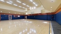 This indoor basketball court is a well-appointed athletic space designed for both recreational and competitive use. The court features a classic, polished hardwood floor with clearly marked court lines, including a regulation three-point arc and key, providing a professional playing surface that reflects the overhead lighting beautifully.

The space is defined by its impressively high ceilings, which accommodate a full-size glass backboard and regulation basketball hoop at one end. The upper portion of the walls is finished in a warm cream tone, while bold navy blue wall padding lines the lower sections, complemented by a rich red accent band in between — a color scheme that adds energy and visual structure to the environment.

Natural light filters in through a row of clerestory windows positioned high along one wall, supplementing the abundant overhead fluorescent strip lighting that ensures consistent, even illumination across the entire court. Observation windows are built into one side wall, allowing spectators to view activity from an adjacent area. The single-floor layout and spacious interior make this court a versatile and functional athletic facility.