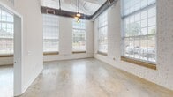 Located at 705 Broadway St in Seneca, South Carolina, this single-floor unit showcases a striking industrial-chic aesthetic rooted in its converted building origins. Soaring ceilings with exposed ductwork and structural columns define the open living and kitchen area, creating a loft-style atmosphere that is both spacious and character-rich. Polished concrete floors run throughout the space, lending a sleek, modern continuity from room to room.

The open-concept living area is bathed in natural light from oversized windows framed by painted white brick walls, offering views of the surrounding streetscape. Pendant Edison bulb lighting hangs from the ceiling, adding warmth and an industrial flair to the space.

The kitchen is well-appointed with dark espresso cabinetry extending to the full height of the tall ceilings, a large white island with a built-in sink, and black stainless steel appliances including a refrigerator, electric range, and over-the-range microwave. The layout promotes easy flow between the kitchen and living areas.

The bathroom features a dark wood vanity with a light countertop, a walk-in shower, and polished tile flooring that complements the concrete found elsewhere. Additional rooms offer generous wall space and access to natural light through large windows. The overall ambiance of this space is open, airy, and distinctly urban, ideal for those who appreciate modern industrial design.
