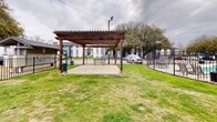 This outdoor community amenity space at 9200 N Normandale St in Fort Worth, Texas, offers a well-maintained gathering area designed for relaxation and recreation. The centerpiece of the space is a sturdy, dark-stained wood pergola structure, supported by four posts and covering a concrete patio pad. The pergola provides a shaded seating area and includes a picnic table beneath it, making it a practical spot for outdoor dining or casual gatherings. Surrounding the pergola is a generous expanse of open green lawn, providing ample room for outdoor activities. The grounds are enclosed by a black iron fence that runs the perimeter of the space, offering both security and a defined boundary. A standout feature visible from the lawn area is a fenced swimming pool, finished in a classic blue tone and surrounded by additional iron fencing for safety. Mature trees are scattered throughout the property, offering natural shade and adding to the greenery of the outdoor environment. The residential buildings of the surrounding community are visible in the background, providing context to the communal nature of this outdoor space. Overall, this amenity area is a functional and accessible outdoor retreat within a residential community setting in Fort Worth.