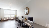 This thoughtfully designed single-floor business center combines a functional workspace with a welcoming lounge area, making it well-suited for both focused work and casual meetings. The workstation zone features a long, sleek gray desk accommodating multiple computer workstations, paired with ergonomic rolling chairs. A distinctive round wooden quatrefoil mirror serves as a standout decorative accent on the crisp white wall above the desk, adding warmth and character to the otherwise clean, contemporary aesthetic. Framed artwork lines the corridor wall, contributing to a polished, professional atmosphere. Rich hardwood-style flooring runs throughout, grounding the space with a warm, sophisticated tone. A glazed gray door connects the workstation area to the adjacent lounge, which is bathed in natural light from a large window fitted with white horizontal blinds. The lounge is furnished with a pair of elegant wood-framed armchairs accented with geometric throw pillows, flanked by round copper-toned side tables — an inviting arrangement ideal for informal conversations or a moment of relaxation. The window offers views of surrounding buildings, reinforcing the urban setting of this well-appointed space.