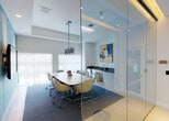 Located at 2263 S Douglas Rd in Miami, FL, this polished single-floor office space delivers a refined, contemporary work environment with thoughtful design details throughout. The layout is anchored by a glass-walled conference room that offers full visual transparency while maintaining an enclosed, professional setting. Inside, a large marble-top conference table is surrounded by modern ergonomic chairs and complemented by a striking linear pendant chandelier. A built-in credenza with a dark countertop, integrated sink, and under-counter appliances lines one wall, adding functional elegance. A vibrant abstract artwork serves as the focal point of the room. Adjacent to the conference room, a secondary meeting or presentation space features a distinctive teal textured accent wall with a wall-mounted display screen and a marble-surface table, accessible through a frameless glass sliding door. The open work area includes built-in granite-topped desks accommodating multiple workstations, blue commercial carpeting underfoot, and clean white walls adorned with botanical framed prints. Recessed lighting and tray ceiling details throughout contribute to the bright, professional ambiance of this well-appointed Miami office.