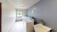 This single-floor shared laundry facility is designed with practicality and functionality in mind. The room is finished with a clean, two-tone color scheme, featuring a muted blue-gray accent wall that contrasts with the lighter surrounding walls, creating a tidy and organized atmosphere. The flooring consists of neutral tile, making maintenance straightforward and easy.

The laundry room is equipped with a row of commercial-grade top-loading washing machines and a matching set of dryers, providing ample capacity for multiple users. A deep utility sink with wall-mounted faucets is conveniently positioned along the accent wall, offering a practical solution for hand-washing or pre-treating garments. A bulletin board is mounted near the entry doors, serving as an informational hub for notices and instructions.

Natural light enters the room through a window fitted with blinds, offering views of the lush greenery and landscaping outside. A baseboard heating unit runs along the exterior wall, ensuring the space remains comfortable year-round. Overhead, a textured ceiling with a centrally mounted flush light fixture provides adequate illumination throughout the room. Two interior doors provide access points, contributing to smooth traffic flow within the facility. Overall, this laundry space is well-appointed and efficiently organized for shared use.