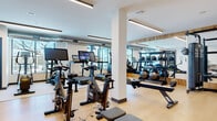 This modern fitness center offers a comprehensive workout environment spread across a single, open floor plan. The space features light hardwood-style flooring throughout, crisp white walls, and recessed linear LED lighting that creates a clean, energizing atmosphere. Structural columns divide the floor into distinct functional zones without disrupting the overall sense of openness.

The cardio zone is well-appointed with multiple treadmills, elliptical trainers, stationary spin bikes equipped with digital display screens, and rowing machines. Large windows along one wall flood this area with natural light and provide views of the surrounding outdoor environment, making workouts feel less confined. Exposed ductwork near the ceiling adds an industrial touch that contrasts effectively with the otherwise polished interior.

The strength training area features a cable machine system, adjustable weight benches, and a well-stocked rack of medicine balls and free weights. Warm wood-panel wall accents frame this section, complemented by floor-to-ceiling mirrors that visually expand the space and allow users to monitor their form. A textured tile accent wall near the entrance adds another layer of visual interest.

Together, these elements combine to create a fully functional, aesthetically refined fitness facility that balances performance and design.