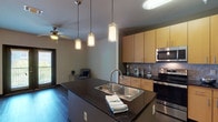 This well-appointed single-floor residence showcases a thoughtfully designed open-concept layout that seamlessly connects the kitchen, dining, and living areas. The kitchen serves as the centerpiece, featuring a large island with dark granite countertops and a stainless steel undermount sink, illuminated by a trio of elegant cylindrical pendant lights. Warm-toned flat-front cabinetry extends to the ceiling, providing generous storage, while a slate-style tile backsplash adds texture and contrast. The full suite of stainless steel appliances includes a refrigerator, range, and built-in microwave. Dark hardwood-style flooring runs throughout the main living areas, complemented by track lighting that adds a contemporary touch. The living area benefits from natural light through a sliding glass door, and a ceiling fan enhances comfort. The bedroom offers plush carpeting and a large window with views of surrounding greenery, filling the room with natural light. A dedicated laundry area with in-unit washer and dryer connections adds everyday convenience. The bathroom is finished with light tile flooring, a soaking tub with tiled surround, and matching granite countertops with wood-toned cabinetry, maintaining a cohesive aesthetic throughout the space.