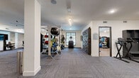 This well-appointed fitness center offers a generous, single-level layout thoughtfully divided into multiple training zones to accommodate a wide range of workout styles. The space features neutral white walls, patterned carpet tile flooring throughout the main areas, and a dedicated room with warm hardwood-style flooring for cycling and group fitness activities.

The cardio zone is lined with large windows that invite natural light and outdoor views, and is outfitted with treadmills, stationary bikes, and elliptical machines, complemented by a large ceiling fan for ventilation. Adjacent to this is a strength training area featuring power racks, cable machines, a full dumbbell rack, and weight benches, all set against floor-to-ceiling mirrors that enhance the sense of space.

A functional training zone includes TRX suspension systems, medicine ball racks, battle ropes, a speed sled track, and a textured climbing wall for dynamic, full-body workouts. Track lighting and recessed fixtures throughout ensure bright, even illumination across all zones.

A separate studio room with hardwood-style flooring, a wall-mounted TV, large mirrors, and cycling bikes provides a dedicated space for group classes or individual cardio sessions. The facility also includes storage shelving and a workstation area, making it a comprehensive and professionally designed fitness destination.