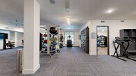This well-appointed fitness center offers a generous, single-level layout thoughtfully divided into multiple training zones to accommodate a wide range of workout styles. The space features neutral white walls, patterned carpet tile flooring throughout the main areas, and a dedicated room with warm hardwood-style flooring for cycling and group fitness activities.

The cardio zone is lined with large windows that invite natural light and outdoor views, and is outfitted with treadmills, stationary bikes, and elliptical machines, complemented by a large ceiling fan for ventilation. Adjacent to this is a strength training area featuring power racks, cable machines, a full dumbbell rack, and weight benches, all set against floor-to-ceiling mirrors that enhance the sense of space.

A functional training zone includes TRX suspension systems, medicine ball racks, battle ropes, a speed sled track, and a textured climbing wall for dynamic, full-body workouts. Track lighting and recessed fixtures throughout ensure bright, even illumination across all zones.

A separate studio room with hardwood-style flooring, a wall-mounted TV, large mirrors, and cycling bikes provides a dedicated space for group classes or individual cardio sessions. The facility also includes storage shelving and a workstation area, making it a comprehensive and professionally designed fitness destination.