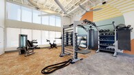 This modern fitness center is thoughtfully designed to support a comprehensive workout experience. The space boasts soaring ceilings with exposed structural elements and large ceiling fans that promote air circulation throughout. Floor-to-ceiling windows along multiple walls flood the interior with generous natural light, creating an open and energizing atmosphere.

The flooring features a durable, textured surface suited for high-traffic exercise use. A full wall of mirrors enhances the sense of space while allowing users to monitor their form during workouts. A striking geometric mural — featuring a retro-inspired palette of orange, teal, navy, gold, and cream — serves as a vibrant focal point and adds a dynamic visual character to the space.

Equipment is diverse and well-organized. Cardio offerings include multiple treadmills and stationary spin bikes, positioned near the windowed walls for a bright workout environment. Strength training is supported by a functional trainer, a multi-use rig with battle ropes and a punching bag, selectorized weight machines, adjustable benches, and a full rack of kettlebells and dumbbells. A step platform rounds out the functional fitness options. The layout provides ample open floor space for stretching or bodyweight exercises, making this fitness center a fully capable facility for users of all training levels.