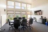 This single-floor conference room is designed to facilitate productive meetings in a well-appointed, light-filled environment. The space is defined by its striking double-height ceilings, which lend an open, airy quality rarely found in standard meeting rooms. A sweeping bank of large windows, dressed with neutral roller shades, lines one wall from floor to ceiling, flooding the interior with abundant natural light and offering views of an outdoor pool area and surrounding palm trees.

The central focus of the room is a substantial oval conference table with a bold red base, surrounded by ergonomic black leather rolling chairs that comfortably seat a full group. A wall-mounted flat-screen television is positioned for easy viewing by all attendees, while a large whiteboard on the adjacent wall supports presentations and collaborative work sessions. Wall sconces provide supplemental lighting, contributing to the room's polished yet functional atmosphere.

A dedicated refreshment station in the corner includes a coffee maker and printer, adding convenience for extended meetings. Entry is provided through a set of glass-paneled double doors, and the carpeted flooring throughout contributes to the room's professional ambiance. Additional seating along the windowed wall accommodates observers or breakout participants.