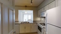 This single-floor space centers around a well-appointed kitchen designed for both functionality and style. The kitchen features white shaker-style cabinetry along the upper and lower sections, providing ample storage throughout. Granite-style countertops in a speckled neutral tone run along the perimeter, complementing the mosaic tile backsplash installed along the cooking wall. A double-basin stainless steel sink is positioned beneath a pass-through window, which opens directly into an adjacent living area, creating a sense of connectivity between the spaces. The living area beyond the pass-through receives natural light through a window dressed with vertical blinds. White appliances, including a freestanding electric range, built-in microwave, refrigerator, and dishwasher, offer a cohesive, clean aesthetic throughout the kitchen. Lighting is provided by a combination of a track-mounted spotlight and a pendant light that hangs through the pass-through opening, adding visual interest. The overall ambiance is bright, clean, and practical, with a neutral color palette that lends a timeless, move-in-ready quality to the space.