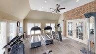 This fitness center offers a thoughtfully designed workout environment housed within a single-floor space distinguished by vaulted ceilings and an abundance of natural light. The architectural character of the room is enhanced by an exposed brick accent wall, which adds warmth and texture alongside the neutral, light-toned walls and striped low-pile carpeting throughout.

The space is well-appointed with a comprehensive selection of exercise equipment. Cardio options include multiple Precor treadmills and an elliptical trainer, while strength training needs are addressed by an Inspire cable/pulley multi-functional machine and a full rack of hex dumbbells in varying weights. A water cooler and wall-mounted television provide added convenience for users during their workouts.

Large windows fitted with horizontal blinds line the walls, offering pleasant views of an outdoor swimming pool and the surrounding grounds, creating an inviting atmosphere. A ceiling fan with integrated lighting ensures comfortable air circulation throughout the room. Full-length mirrors along one wall visually expand the space and support proper form during exercise. French doors provide access to the exterior, further connecting the fitness center to the outdoor amenities. Recessed lighting complements the natural light, keeping the space bright and functional at all hours.