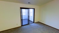 This single-floor space offers a clean, functional layout with a straightforward design. The main room features wall-to-wall carpeting in a neutral tone, providing a comfortable and quiet underfoot feel throughout. Smooth, cream-colored walls and standard baseboards create a simple, understated aesthetic that lends itself well to a variety of uses and furnishing styles.

A standout feature of the space is the large black-framed sliding glass door, which opens into an adjacent enclosed room — likely a sunroom or covered patio area. This secondary space benefits from multiple windows that allow natural light to filter through, brightening the interior. The overhead HVAC vent in the ceiling ensures year-round climate control throughout the space.

The overall ambiance is modest and practical, presenting a blank-canvas quality that makes it highly adaptable. Whether used as a residential living space or a small office, the layout's simplicity and the added bonus of the enclosed sunroom-style area provide both versatility and a touch of openness. The connection between the interior room and the windowed enclosure creates a layered sense of space and allows for passive natural light even when the sliding door is closed.