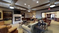 This inviting lounge space exudes a classic, lodge-inspired aesthetic defined by warm tones, rich wood detailing, and carefully chosen furnishings. The focal point of the room is a stacked-stone fireplace, which anchors the seating area and adds a rustic yet refined character to the space. Above the fireplace, a flat-screen television is mounted within a built-in wood surround, flanked by shelving that adds both functionality and visual depth.

The ceiling features a coffered design with recessed lighting and pendant fixtures, lending a sense of architectural structure while maintaining a warm, welcoming ambiance. Dark wood wainscoting lines the lower walls throughout, complementing the earthy beige and brown color palette of the space.

Seating is arranged thoughtfully around a pair of dark wood coffee tables, with plush sofas and upholstered armchairs providing ample room for gathering. Framed artwork adorns the walls, adding decorative interest alongside potted greenery that introduces a natural element. The patterned carpet grounds the room and ties together its cohesive, comfortable design. Overall, the space offers a relaxed yet polished atmosphere well-suited for socializing or unwinding.