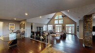 This single-story space makes a bold architectural statement through its dramatic vaulted ceiling, which peaks at a striking height in the main living area. A large arched clerestory window crowns the roofline, flooding the interior with natural light and drawing the eye upward. The open-concept living area is expansive, featuring rich dark hardwood flooring that runs throughout and neutral gray walls that complement the modern aesthetic. Multiple sets of French doors line the exterior walls, offering seamless access to the outdoors while maximizing natural light and views of the surrounding greenery.

The living area is defined by its generous proportions, accommodating multiple seating arrangements beneath the sweep of the vaulted ceiling. A sleek ceiling fan anchors the upper space, adding both function and visual interest. A stacked stone column accent provides an earthy, textural contrast to the otherwise contemporary finishes. The transition to a lower level is marked by a modern cable-rail railing with a stone-tiled landing, adding architectural detail.

The kitchen features a galley-style layout with dark espresso cabinetry, granite countertops, a mosaic tile backsplash, stainless steel appliances, and track lighting, delivering a polished and functional culinary space that complements the overall modern character of the home.