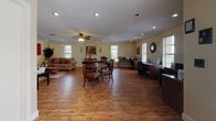 This single-story leasing office is thoughtfully designed to provide a functional and welcoming environment for residents and visitors alike. The space is finished throughout with warm hardwood-style flooring, crown molding, and recessed lighting, lending a polished, cohesive aesthetic across all areas.

Guests are greeted upon entry by a dedicated reception foyer, complete with a ceiling fan, accent seating — including a red leather chair and a brown armchair — decorative artwork, and a "Welcome" sign that sets a hospitable tone. A patterned area rug bearing the "Leasing Office" designation marks the entryway clearly.

The main open area is generously proportioned and multi-functional, accommodating a central dining-style table with chairs, a full lounge seating arrangement with two sofas, red accent pillows, a glass coffee table, and a wall-mounted flat-screen television flanked by bold red artwork. Ceiling fans circulate air throughout, and multiple windows with slatted blinds allow natural light to fill the space.

A dedicated business center lines one wall, featuring multiple computer workstations set along a continuous built-in desk, office chairs, and windows overlooking greenery outside. Decorative wall clocks and colorful artwork complete the area, creating a productive yet inviting workspace for residents.