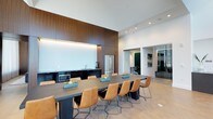 This single-floor space offers a sleek, contemporary design highlighted by soaring ceilings that amplify the sense of openness and airiness throughout. The central feature is a generous dark-toned dining or conference table surrounded by tan leather chairs with metal sled bases, creating a refined yet approachable setting suitable for both social gatherings and professional meetings.

Along one wall, a built-in kitchenette is seamlessly integrated behind rich, dark wood panel cabinetry that extends floor to ceiling, providing a striking architectural focal point. The kitchenette is equipped with a stainless steel refrigerator, microwave, and a sink, offering convenient functionality. Recessed under-cabinet lighting adds a warm, ambient glow against the clean white backsplash.

The space is finished with light hardwood flooring that contrasts elegantly with the darker furnishings and cabinetry. Recessed ceiling lighting ensures even illumination throughout. An adjacent glass-enclosed room is visible through sliding glass doors, offering additional flexible space for private meetings or breakout sessions. The overall ambiance is polished, modern, and highly functional.