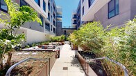 Located at 1650 Lincoln Blvd in Santa Monica, California, this amenity space is centered around a beautifully designed courtyard framed by a modern multi-story building with a clean, contemporary façade of smooth concrete, large blue-tinted windows, and glass-railed balconies. The open-air courtyard serves as the heart of the space, featuring large-format concrete pavers, generous seating arrangements with stylish outdoor furniture, and raised Corten steel planter beds filled with a curated mix of bamboo, snake plants, ornamental grasses, and drought-tolerant succulents that reflect Southern California's landscaping sensibility. Multiple distinct seating zones offer flexible gathering areas, including dining tables with chairs and lounge configurations with cushioned sofas and chairs in muted tones. A partially covered indoor-outdoor transition area connects the courtyard to an interior lounge, where bold orange furnishings—including sofas, chairs, and coffee tables—add a vibrant pop of color against light tile walls and polished concrete-look flooring. Adjacent to the lounge, a fitness area with exercise equipment is visible through glass partitions. String lights are strung overhead in the covered outdoor section, adding warmth and ambiance. The entire space is bathed in abundant natural light, creating a welcoming, resort-like atmosphere ideal for relaxation and community gathering.