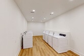 This communal laundry facility at 8701 Old Spanish Trail in Ocean Springs, Mississippi, offers a practical and well-appointed space designed for efficient, shared use. The room is finished with crisp white walls and warm-toned luxury vinyl plank flooring that lends a clean, modern aesthetic throughout. Recessed lighting is evenly distributed across the ceiling, ensuring bright, consistent illumination across the entire space. Multiple top-loading washing machines and dryers are arranged in two parallel rows along the walls, maximizing the usable floor area and allowing for comfortable circulation between machines. The machines are all white, contributing to the room's cohesive and tidy appearance. HVAC vents are integrated into the ceiling, maintaining comfortable climate control year-round. A door with a sidelight window is visible at one end of the room, providing both access and a touch of natural light. The layout is straightforward and functional, with open floor space between machine rows for easy movement. White baseboards frame the room neatly, reinforcing the clean, well-maintained character of the facility. Overall, this laundry room presents a highly functional, orderly environment suited to serve the needs of a multi-unit residential community.