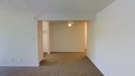This single-floor space features an open layout with carpeted flooring that extends throughout the main living areas. The interior is characterized by neutral beige walls and a textured popcorn ceiling, creating a clean and understated aesthetic. A ceiling fan is centrally mounted, providing both lighting and air circulation to the space. The layout reveals a partially open floor plan, where a structural beam defines the transition between the main living area and an adjoining room, maintaining a sense of connectivity while subtly delineating separate spaces. A closet with shelving is visible in the background, offering built-in storage. The rooms are bathed in soft, ambient light, contributing to a calm and functional atmosphere. The overall design is straightforward and practical, with minimal architectural detailing, making it a versatile space adaptable to various furnishing arrangements and uses.