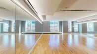 This single-floor fitness or dance studio is designed with both function and aesthetics in mind. The centerpiece of the space is the expansive, polished hardwood floor, which provides a smooth and durable surface well-suited for dance, yoga, or group fitness activities. Large floor-to-ceiling mirrors line multiple walls, visually expanding the room while allowing participants and instructors to monitor form and movement with ease. The mirrors are fitted with integrated linear lighting strips that cast a bright, even illumination throughout the studio.

Natural light is a prominent feature, entering through large windows that offer views of the surrounding urban environment. The windows are framed by textured grey wall panels, lending a refined and contemporary aesthetic to the space. The ceiling is finished in a clean white with subtle tiered architectural detailing, adding depth without visual clutter.

Additional features include built-in storage with white cabinet doors along one wall, a dark-framed glass door providing access to an adjacent room—visible to contain fitness equipment—and perimeter baseboard heating beneath the windows. The overall ambiance is clean, professional, and well-appointed, making it a versatile and welcoming environment for a wide range of physical activities.