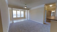 This single-floor space features an open layout that combines a carpeted main living area with a tiled windowed alcove, creating a functional and versatile floor plan. The two areas are elegantly divided by decorative columns, adding architectural character while maintaining a sense of openness throughout.

The main living area is finished with neutral-toned carpet and features crown molding along the ceiling perimeter, lending a polished, traditional aesthetic. A ceiling fan with an integrated light fixture provides both ventilation and ambient lighting. The walls are painted in a warm, neutral beige tone, complemented by crisp white baseboards that run throughout.

The adjacent windowed alcove is finished with tile flooring and benefits from multiple windows that allow natural light to fill the space. A second ceiling fan mirrors the one in the main area, ensuring consistent comfort throughout. The large windows offer views to the exterior and contribute significantly to the bright, airy ambiance of the space.

Overall, the space presents a clean, well-maintained interior with consistent finishes, practical features, and a neutral palette that provides a versatile foundation for a variety of furniture arrangements and design preferences.