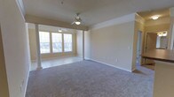 This single-floor space features an open layout that combines a carpeted main living area with a tiled windowed alcove, creating a functional and versatile floor plan. The two areas are elegantly divided by decorative columns, adding architectural character while maintaining a sense of openness throughout.

The main living area is finished with neutral-toned carpet and features crown molding along the ceiling perimeter, lending a polished, traditional aesthetic. A ceiling fan with an integrated light fixture provides both ventilation and ambient lighting. The walls are painted in a warm, neutral beige tone, complemented by crisp white baseboards that run throughout.

The adjacent windowed alcove is finished with tile flooring and benefits from multiple windows that allow natural light to fill the space. A second ceiling fan mirrors the one in the main area, ensuring consistent comfort throughout. The large windows offer views to the exterior and contribute significantly to the bright, airy ambiance of the space.

Overall, the space presents a clean, well-maintained interior with consistent finishes, practical features, and a neutral palette that provides a versatile foundation for a variety of furniture arrangements and design preferences.