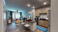 This well-appointed single-floor residence offers a thoughtfully designed open-concept layout that seamlessly connects the kitchen, dining, and living areas. The kitchen is a standout feature, showcasing light wood-grain cabinetry extending to the ceiling, dark countertops, a full suite of stainless steel appliances including a refrigerator, range, and microwave, and a central island with bar seating — ideal for casual dining and entertaining. Wood-look flooring flows throughout the common areas, while recessed lighting and a ceiling fan contribute to a bright, comfortable ambiance. Large windows allow generous natural light into the living room, which is anchored by a teal accent wall and access to the entry foyer. The primary bedroom offers plush carpeting, a ceiling fan, and a large window, leading into a well-designed en-suite bathroom with dual vanities, wood-tone cabinetry, dark countertops, and direct access to a generously sized walk-in closet with multi-tier shelving and hanging rods. A second bedroom features its own bold blue accent wall and ample natural light. The second bathroom includes a tub-shower combination with wood-look flooring, completing this stylish and functional residence.